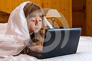 Cute little girl using a laptop in bed under a blanket