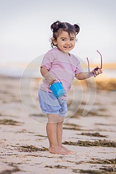 Cute little girl walking on sandy beach, holding bucket and sunglasses. Warm day. Happy childhood. Summer vacation. Holiday