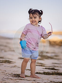 Cute little girl walking on sandy beach, holding bucket and sunglasses. Warm day. Happy childhood. Summer vacation. Holiday