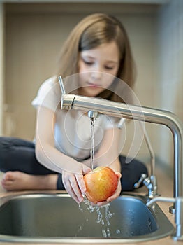 A cute little girl washes a red yellow apple in a sink. Child is washing the fruit in kitchen. Little child learns the rules