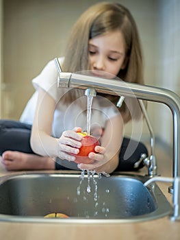 A cute little girl washes a red yellow apple in a sink. Child is washing the fruit in kitchen. Little child learns the rules