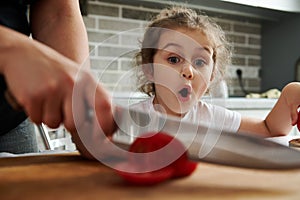 Beautiful little girl watches her mother cutting tomato on a cutting board. Mother and daughter preparing food together