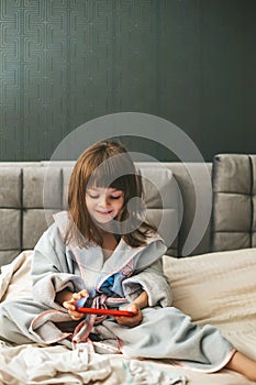 A cute little girl wearing a bathrobe sitting on a bed, completely engrossed in her smartphone