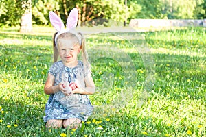 Cute little girl wearing bunny ears on Easter day. Girl sitting on a grass and holding painted Easter eggs