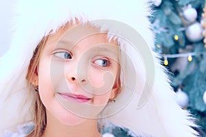 Cute little girl wearing a white fluffy hat looking to the side over Christmas tree background.
