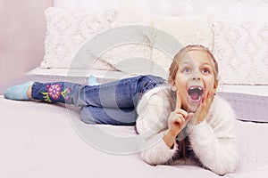 Cute little girl wearing a white sweater and blue jeans lying on the bad and gesturing.