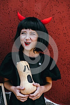 Cute little girl  wearing wig with red horns holding pumpkin on a background of red wall. Halloween celebration party portrait