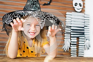 Cute little girl wearing witch hat sitting behind a table in Halloween theme decorated living room, making scary face.