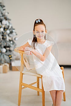 Cute little girl in white dress sitting on a chair by Christmas tree.