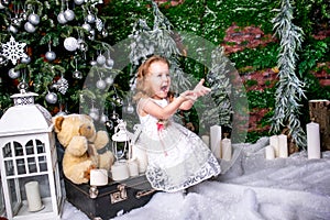 Cute little girl in a white dress sitting near a Christmas tree on a suitcase next to the candles and a teddy bear, throws snow up