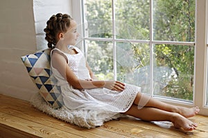 Cute little girl in white dress with two ponytails sitting at window sill looking at the street