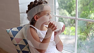 Cute little girl in white dress with two ponytails sitting at window sill looking at the street and drinking hot tea from white cu