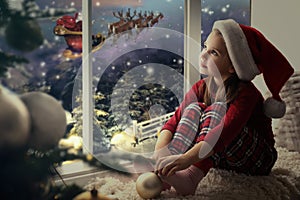 Cute little girl on window sill at home waiting for Santa Claus. Christmas celebration