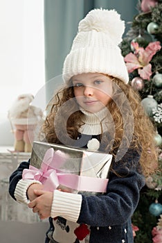 Cute little girl in winter clothes posing in home front of Christmas tree with gift box in hand