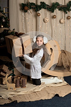 Cute little girl in winter clothes posing in home front of Christmas tree with gift box in hand