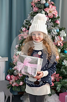 Cute little girl in winter clothes posing in home front of Christmas tree with gift box in hand