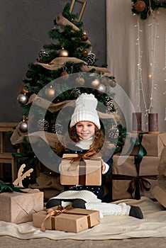 Cute little girl in winter clothes posing in home front of Christmas tree with gift box in hand