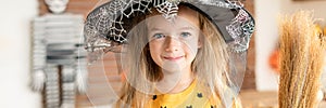 Cute little girl in witch costume holding a broom is standing in Halloween decorated living room, looking at camera and smiling.