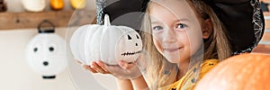 Cute little girl in witch costume holding hand painted pumpkin and smiling. Halloween concept.