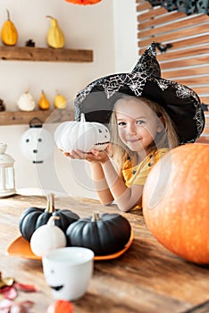 Cute little girl in witch costume sitting behind a table in Halloween theme decorated room, holding hand painted pumpkin.