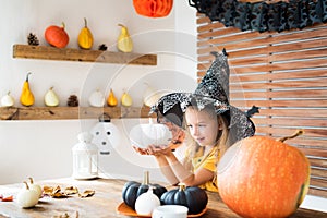 Cute little girl in witch costume sitting behind a table in Halloween theme decorated room, holding hand painted pumpkins smiling.