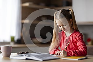 Cute Little Girl Writing In Notepad While Sitting At Desk At Home