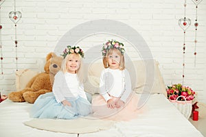 Cute little girls in dresses with flowers wreath on their head. Two little sisters sitting on the bed in white studio.