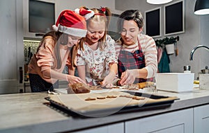 Cute little girls in red Santa hats with mother making homemade dough Christmas gingerbread cookies using cookie cutters together