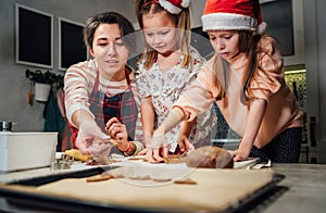 Cute little girls in red Santa hats with mother making homemade dough Christmas gingerbread cookies using cookie cutters together