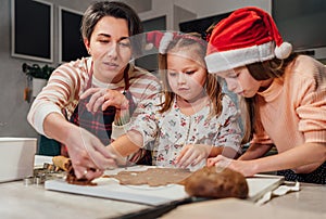 Cute little girls in red Santa hats with mother making homemade dough Christmas gingerbread cookies using cookie cutters together