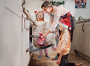 Cute little girls in red Santa hats with mother making homemade sweet biscuits. They putting baking sheet in hot oven. Christmas