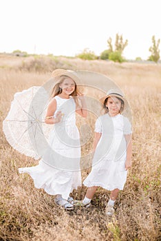 Cute little girls sisters with blond  hair in a summer field at sunset in white dresses