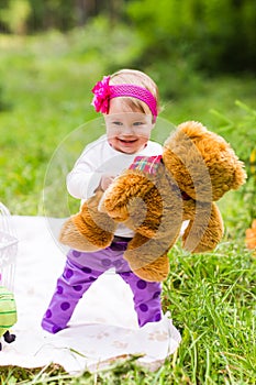 Cute little happy baby girl with big brown teddy bear on green grass meadow, spring or summer season
