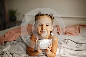 Cute little happy girl with two ponytails in her bed drinking hot tea from white cup