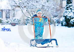 Cute little kid boy in colorful winter clothes having fun with snow shovel, outdoors during snowfall. Active outdoors
