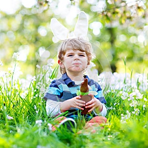 Cute little kid boy with Easter bunny ears celebrating traditional feast. Happy child eating chocolate rabbit fugure on