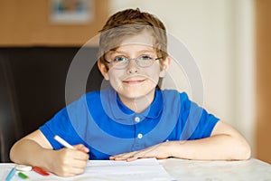 Cute little kid boy with glasses at home making homework, writing letters with colorful pens.