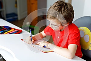 Cute little kid boy with glasses at home making homework, writing letters with colorful pens.