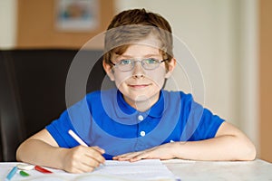 Cute little kid boy with glasses at home making homework, writing letters with colorful pens.