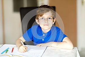 Cute little kid boy with glasses at home making homework, writing letters with colorful pens.