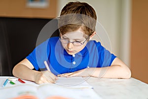 Cute little kid boy with glasses at home making homework, writing letters with colorful pens.