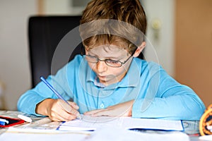 Cute little kid boy with glasses at home making homework, writing letters and doing maths with colorful pens. Little