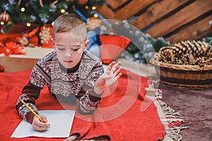 Cute little kid in festive dress lying on the floor, writing letter to Santa. Christmas tree with lights and gifts at photo