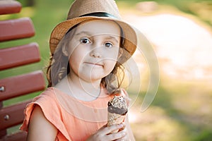 Cute little kid girl in a hat eating ice cream cone. Child sitting on a park bench and laughing . Summer concept.