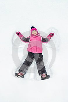 Cute little kid girl in warm ski sport suit making snow angel outdoors. Kid having fun lying on snowdrift after snow