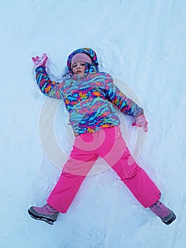 Cute little kid girl in warm ski sport suit making snow angel outdoors. Kid having fun lying on snowdrift after snow