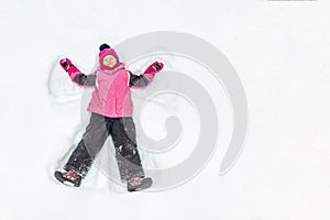Cute little kid girl in warm ski sport suit making snow angel outdoors. Kid having fun lying on snowdrift after snow storm in