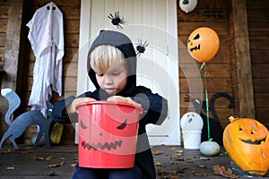 Cute little kid in halloween costume sitting on porch of house, holding basket and looking at how much candy he collected while