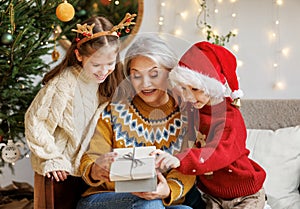 Little children granddaughter and grandson giving Christmas gift box to smiling grandmother during winter holidays