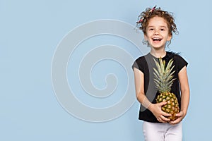 Cute little laught girl holding a pineapple, over blue background, studio shoot. Space for text.